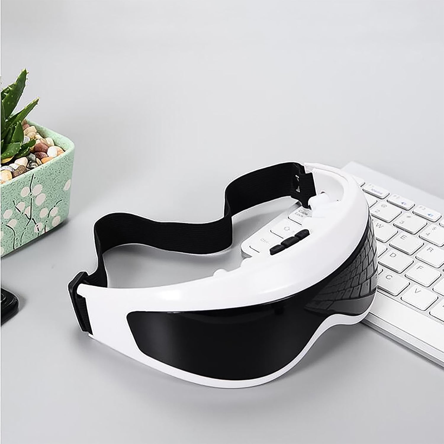 white and black eye massager with adjustable strap on desk near keyboard and plant best eye massager for relaxation